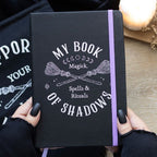 Cuaderno negro con texto "My Book of Shadows" y dibujo de escobas blancas, con goma elástica y cinta de lectura morada.