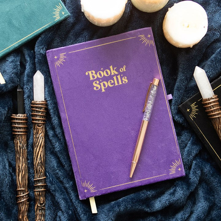 Cuaderno morado de terciopelo con el texto "Book of Spells" en dorado junto a un bolígrafo de amatista rodeado de velas blancas.