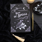 Cuaderno negro con ilustración blanca de un cuervo sobre una rama, lunas y el texto "Book of Spells" con una borla dorada.