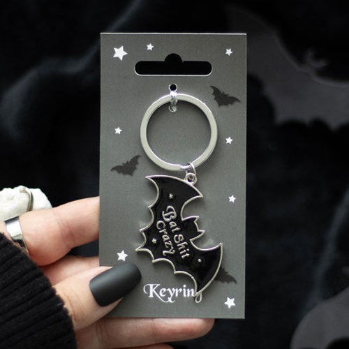 Llavero de murciélago negro con texto plateado "Bat Shit Crazy" sostenido por una mano con uñas negras.