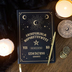 Cuaderno negro con diseño dorado de tablero Ouija, alfabeto, lunas y estrellas sobre fondo blanco.